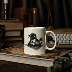 Puede incluir: Una taza de cer&aacute;mica blanca con una ilustraci&oacute;n en tinta negra de una criatura con cuernos leyendo un libro. La taza tiene un cuerpo cil&iacute;ndrico liso con un asa curva en el lado derecho. El gr&aacute;fico est&aacute; centrado en la parte frontal de la taza.