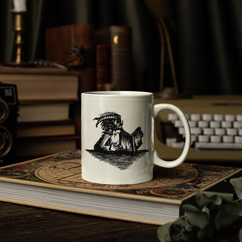 Puede incluir: Una taza de cer&aacute;mica blanca con una ilustraci&oacute;n en tinta negra de una criatura con cuernos leyendo un libro. La taza tiene un cuerpo cil&iacute;ndrico liso con un asa curva en el lado derecho. El gr&aacute;fico est&aacute; centrado en la parte frontal de la taza.