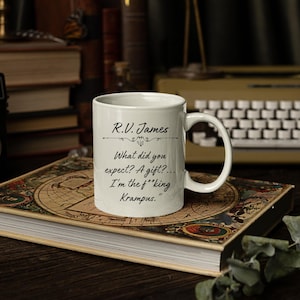 Puede incluir: Una taza de cer&aacute;mica blanca con texto impreso en negro que dice: R.V. James. What did you expect? A gift?... I'm the f**king Krampus. La taza tiene un asa est&aacute;ndar y un cuerpo cil&iacute;ndrico liso con un acabado brillante y letras claras y legibles.
