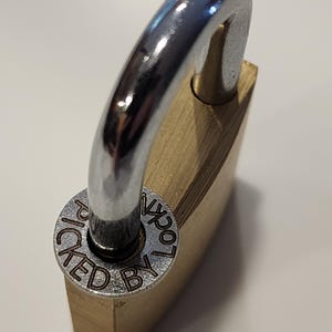 May include: A brass padlock with a shiny silver shackle. The circular base of the shackle reads "PICKED BY LOCKN". The lock is set against a white background, highlighting its metallic finish and design.