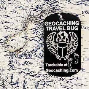 Op de afbeelding: Zwarte hondenpenning met de tekst "GEOCACHING TRAVEL BUG" en een scarabee-ontwerp met vleugels. De penning bevat ook de tekst "Trackable at Geocaching.com" en twee voetafdrukken. Een zilveren kogelketting is bevestigd.