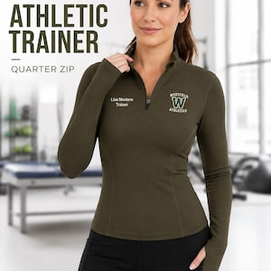 May include: An olive green long-sleeve quarter-zip athletic top with a high collar. The chest features the text 'Lisa Montero Trainer' and a 'Westfield Athletics' logo with a large letter W. The top has thumb holes at the sleeve cuffs.