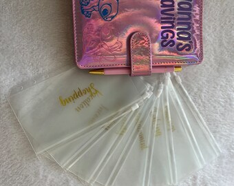 May include: A pink, iridescent savings binder with a cartoon character design and the text "Ivanna's Savings". Includes clear, zippered budget envelopes with gold lettering. A pink pen is included.
