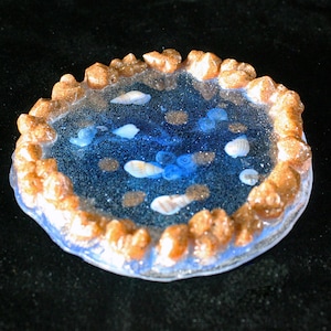 Puede incluir: Un posavasos redondo de vidrio transparente con un centro de resina azul y un borde de resina marrón texturizado. El centro del posavasos presenta una variedad de pequeñas conchas blancas y purpurina azul.