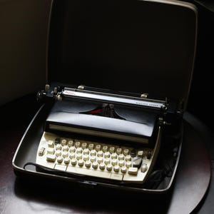 Könnte beinhalten: Eine schwarze Vintage-Schreibmaschine mit einer weißen Tastatur und dem Text "Electric Power" oben. Die Schreibmaschine ist geöffnet und befindet sich in einem schwarzen Transportkoffer.