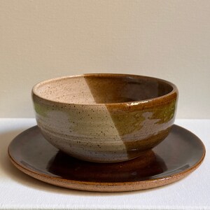 Puede incluir: Un cuenco de cerámica marrón y beige con un esmalte marrón en el exterior y un esmalte beige moteado en el interior. El cuenco está sobre un plato de cerámica marrón a juego.