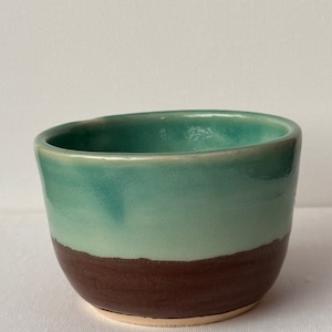 Puede incluir: Un pequeño cuenco redondo de cerámica con un esmalte verde claro en la mitad superior y un esmalte marrón oscuro en la mitad inferior.