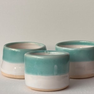 May include: Three ceramic cups with a teal glaze and white rim. The cups are different sizes and are arranged in a row.