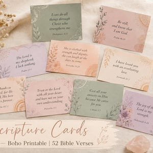 Può includere: Una collezione di cartoline rettangolari con versetti biblici in tonalità pastello verde, rosa, pesca e viola, decorate con illustrazioni floreali. Una penna dorata e fiori secchi sono disposti accanto alle carte su un tessuto.