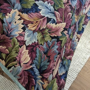 May include: A fabric textile featuring a dense, repeating pattern of stylized leaves in shades of deep burgundy, forest green, muted blue, and cream, set against a black background. The material has a visible woven texture throughout the surface.