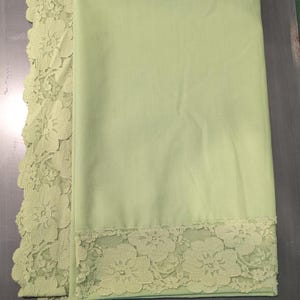 May include: A light sage green fabric textile featuring a smooth, solid-colored central panel bordered by a wide, intricate floral lace trim along one vertical edge and one horizontal edge, showcasing a delicate scalloped edge design.