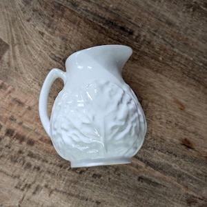 May include: A small white ceramic creamer pitcher featuring a raised, embossed leaf pattern covering the exterior surface. The pitcher has a curved handle on the side and a distinct pouring spout at the top rim, with a glossy, smooth finish.