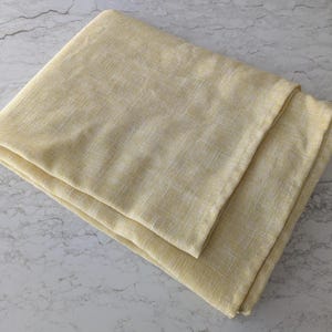 May include: Folded pale yellow linen fabric. The fabric has a subtle woven texture and a soft, muted color. The folded edges create a layered effect, suggesting a tablecloth or other textile.