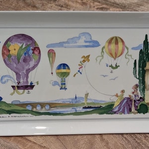 May include: A rectangular white ceramic serving tray with integrated handles, featuring a colorful watercolor-style illustration of hot air balloons, a kite, a stone building, trees, and a bridge over a river against a light background.