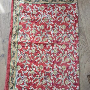 May include: A rectangular textile featuring a terracotta red background with a repeating pattern of light green pinecones and white leafy branches. A wide border in a muted yellow tone displays a dark green botanical vine motif.