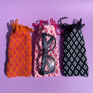 May include: Three knitted eyeglass cases in orange, pink, and navy blue, each with a diamond pattern. The pink case holds a pair of eyeglasses. The cases have drawstring closures with matching colored cords.