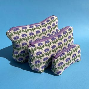 May include: Three knitted zippered pouches in different sizes, patterned with purple flowers and green leaves on a white background. Each pouch has a purple zip, set against a blue backdrop.
