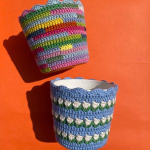 Puede incluir: Dos fundas de ganchillo para macetas. Una es una mezcla de hilos coloridos en tonos rosa, amarillo, azul y verde. La otra es azul con detalles blancos y verdes. Ambas tienen un borde festoneado.