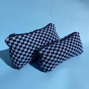 May include: Two zippered pouches with a black and light blue chequered pattern. The rectangular pouches are made of a knitted fabric. The background is a solid light blue.