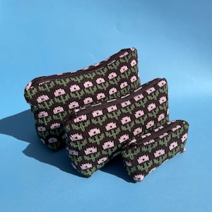 May include: Three knitted zippered pouches in different sizes, patterned with white flowers and green leaves on a black background. The pouches are displayed against a blue backdrop, showcasing their design and texture.
