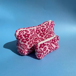 May include: Two knitted pouches in shades of pink and white, with a repeating floral pattern. The larger pouch is rectangular, and the smaller one is a similar shape. Both have white zips. The background is a solid light blue.