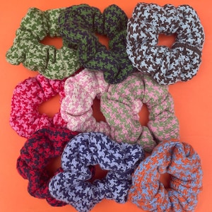 May include: A collection of handmade scrunchies in various colors and patterns. The scrunchies feature a houndstooth design in shades of green, pink, blue, red, and orange. The scrunchies are made of a soft, knitted material and are arranged on an orange background.