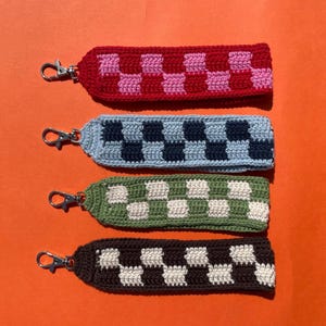 Puede incluir: Cuatro correas de muñeca de ganchillo con cierres plateados. Cada correa presenta un patrón de tablero de ajedrez en diferentes combinaciones de colores: rojo y rosa, azul claro y marino, verde y blanco, y marrón y blanco. Las correas se muestran sobre un fondo naranja.