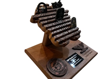 May include: A wooden award with a dark brown base and a multi-tiered, carved design. The award features several small, dark-colored emblems, including a steering wheel, a ship, and a shield. Text on the base reads "Congratulations Captain Select!"