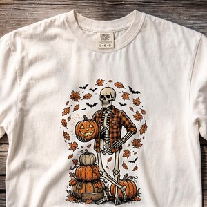 Puede incluir: Una camiseta de cuello redondo color hueso con un gráfico de un esqueleto con camisa de cuadros naranja y negra, sosteniendo una calabaza. El esqueleto está junto a una pila de calabazas, rodeado de hojas de otoño y pequeños murciélagos negros.
