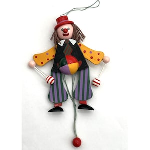 May include: A wooden clown marionette featuring a red hat, brown yarn hair, and a black vest with a striped tie. The clown has yellow sleeves with orange dots, purple and green striped pants, and holds two white and red striped drumsticks.