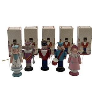 Könnte beinhalten: Fünf hölzerne Nussknacker-Ornamente in einer Reihe, jeweils mit einer eigenen rechteckigen Schachtel. Die Figuren umfassen eine Fee, eine Maus, einen Soldaten in Rot, einen Trommler in Blau und eine Figur in einem rosa Kleid, alle mit goldenen Schnüren.