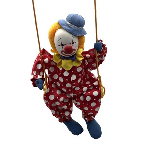 Op de afbeelding: Een vintage clownpop op een schommel. De pop heeft een wit gezicht met geschilderde kenmerken, een rode neus en een blauwe hoed. Ze draagt een rode outfit met witte stippen, een gele kraag en blauwe schoenen. De schommel is van touw.