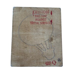May include: A wooden board with a wire outline of a lightbulb shape. The board has the text "CAUTION THIS SIDE AGAINST METAL SURFACE" in red. The board also has the text "This die was inspected and tested by" and a letter "Q".
