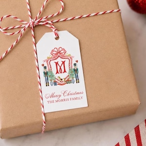 May include: A white rectangular gift tag tied with red and white baker's twine to a brown paper package. The tag features a red letter M, two nutcrackers, a rocking horse, and the text Merry Christmas THE MORRIS FAMILY in red ink.