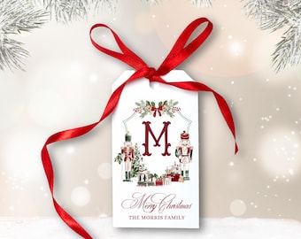 May include: White rectangular Christmas gift tag with a red ribbon. The tag features a large burgundy letter "M", two nutcracker illustrations, and the text "Merry Christmas" and "THE MORRIS FAMILY". The tag is decorated with festive greenery.