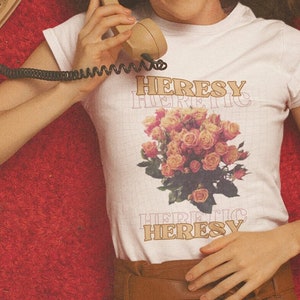 Może przedstawiać: Kobieta w białej koszulce z nadrukiem kwiatowym i tekstem "HERESY HERETIC HERESY". Trzyma do ucha staromodny telefon tarczowy i leży na czerwonym dywanie.