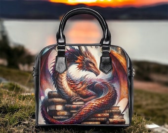 May include: Black leather handbag featuring a detailed illustration of a red and orange dragon resting on a stack of books. The dragon has large wings and is set against a blurred background of a sunset over water. The bag has a top handle.