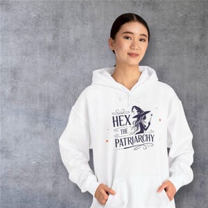 May include: White hoodie with the text "HEX THE PATRIARCHY" in a dark blue font, along with an illustration of a witch. The hoodie has a drawstring and a front pocket.