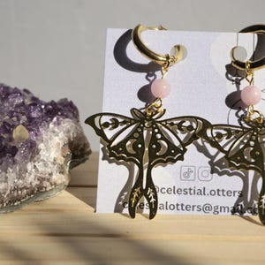 Può includere: Orecchini a forma di falena color oro con un accento di perline rosa. Gli orecchini sono esposti su una carta bianca con informazioni sui social media. Sullo sfondo è visibile un grande cristallo di ametista viola.
