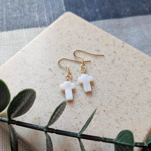May include: A pair of gold earrings with white mother of pearl cross charms. The earrings are on a light brown surface.