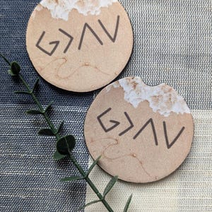 May include: Two round brown car coasters with a white and brown marbled design. The coasters feature the text "God is Greater Than the Highs and Lows" in black.