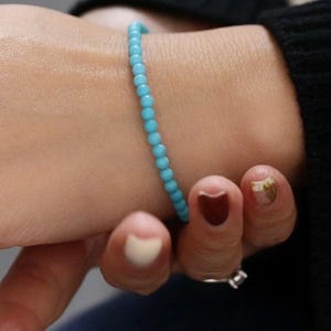 Op de afbeelding: Een delicate armband bestaande uit kleine, ronde, turquoise kralen. De armband wordt om een pols gedragen, met een zwarte mouw op de achtergrond. De hand heeft gelakte nagels, met een ring aan een vinger.