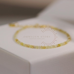 Puede incluir: Una delicada pulsera dorada con pequeñas piedras preciosas amarillas de corte cuadrado. La pulsera se exhibe sobre una superficie blanca. El texto "OHMYGEM STORY HANDMADE JEWELLERY" es visible en el fondo.