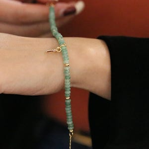 Op de afbeelding: Een delicate armband met kleine, lichtgroene kralen geregen met goudkleurige accenten. Een kleine, goudkleurige sleutelbedel hangt aan de armband. De armband wordt om een pols gedragen, met een zwarte mouw op de achtergrond.