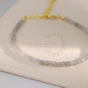 May include: A delicate bracelet featuring small, square-cut, light grey beads strung together. The bracelet has a gold-coloured chain and clasp. The text "OHMYGEM STORY HANDMADE JEWELLERY" is visible on the background.
