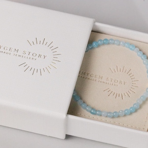 May include: A light blue beaded bracelet is displayed in an open white box. The box is embossed with the text "OHMYGEM STORY HANDMADE JEWELLERY" in gold. The bracelet is made of small, round, light blue beads.