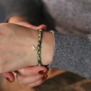 Peut inclure: Un bracelet de perles vertes et noires avec des détails dorés. Il comporte un petit charme doré en forme de lettre "K". Le bracelet est porté au poignet, avec un pull gris en arrière-plan.