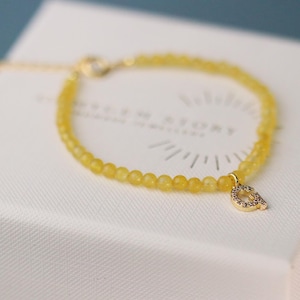 Könnte beinhalten: Ein zartes gelbes Perlenarmband mit einem goldfarbenen Anhänger in Form des Buchstabens "Q", verziert mit kleinen, klaren Steinen. Das Armband liegt auf einer weißen Schachtel mit Logo.