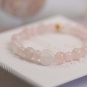 Könnte beinhalten: Ein Armband aus Rosenquarzperlen mit einem herzförmigen Anhänger. Das Armband besteht aus hellrosa, runden Perlen und einer herzförmigen Perle. Ein kleiner goldfarbener Akzent ist sichtbar. Das Armband wird auf einer weißen Oberfläche präsentiert.