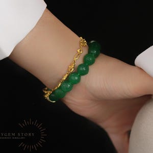 May include: A gold bracelet with a delicate chain and a green beaded bracelet are worn on a wrist. The green beads are faceted and round. The background is black.
